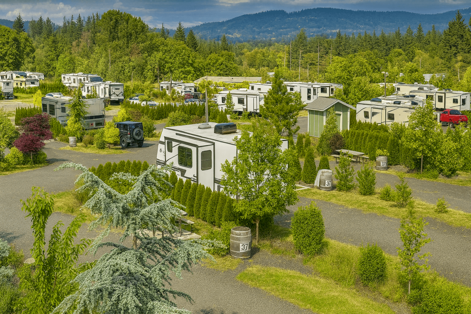A scenic RV park with white trailers and campers among green trees, shrubs, and pathways, set against distant mountains, under a bright, clear sky.