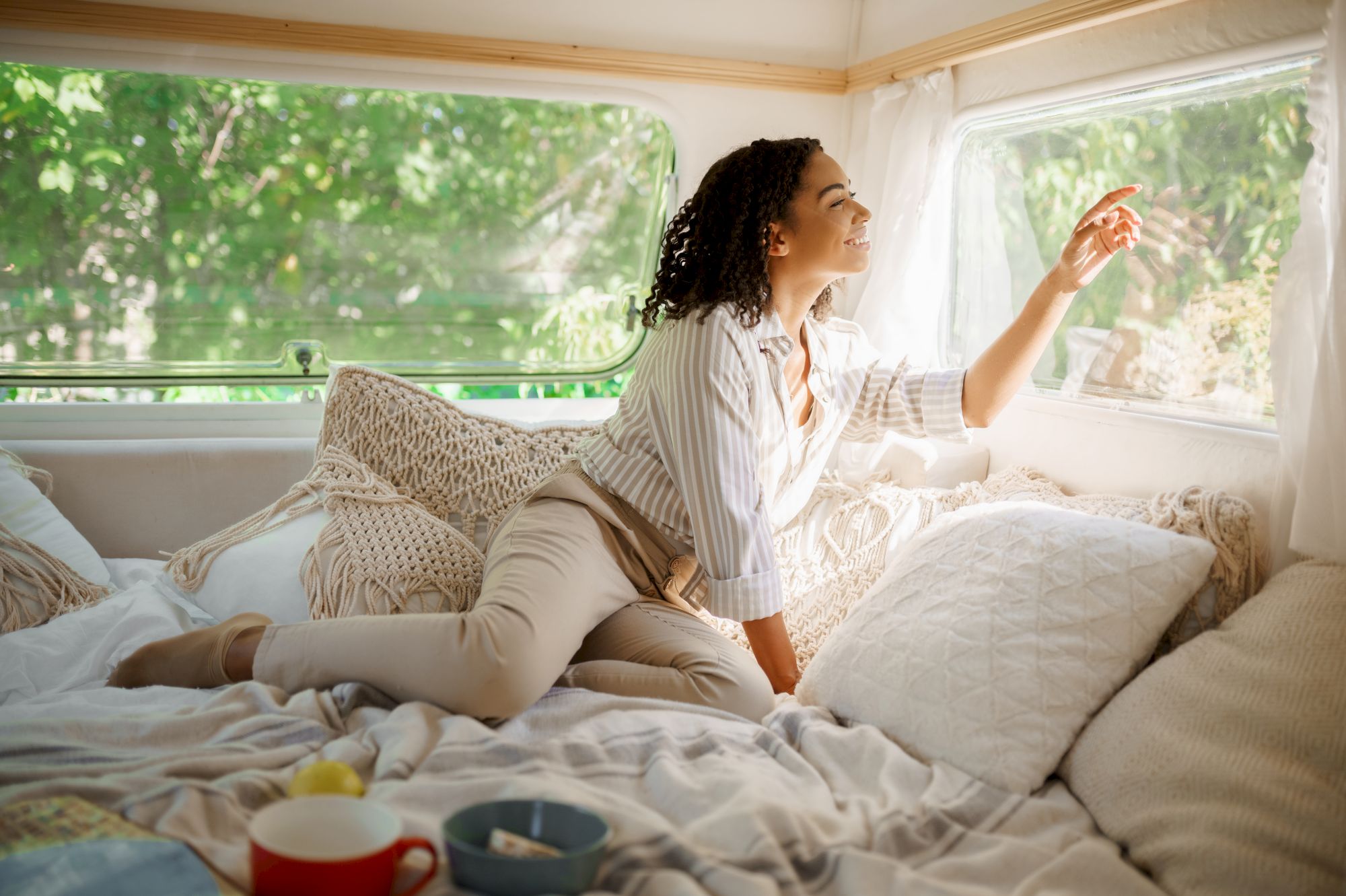 A person enjoys a cozy moment on a bed inside a caravan, looking out the window, surrounded by pillows and a cup on the side.