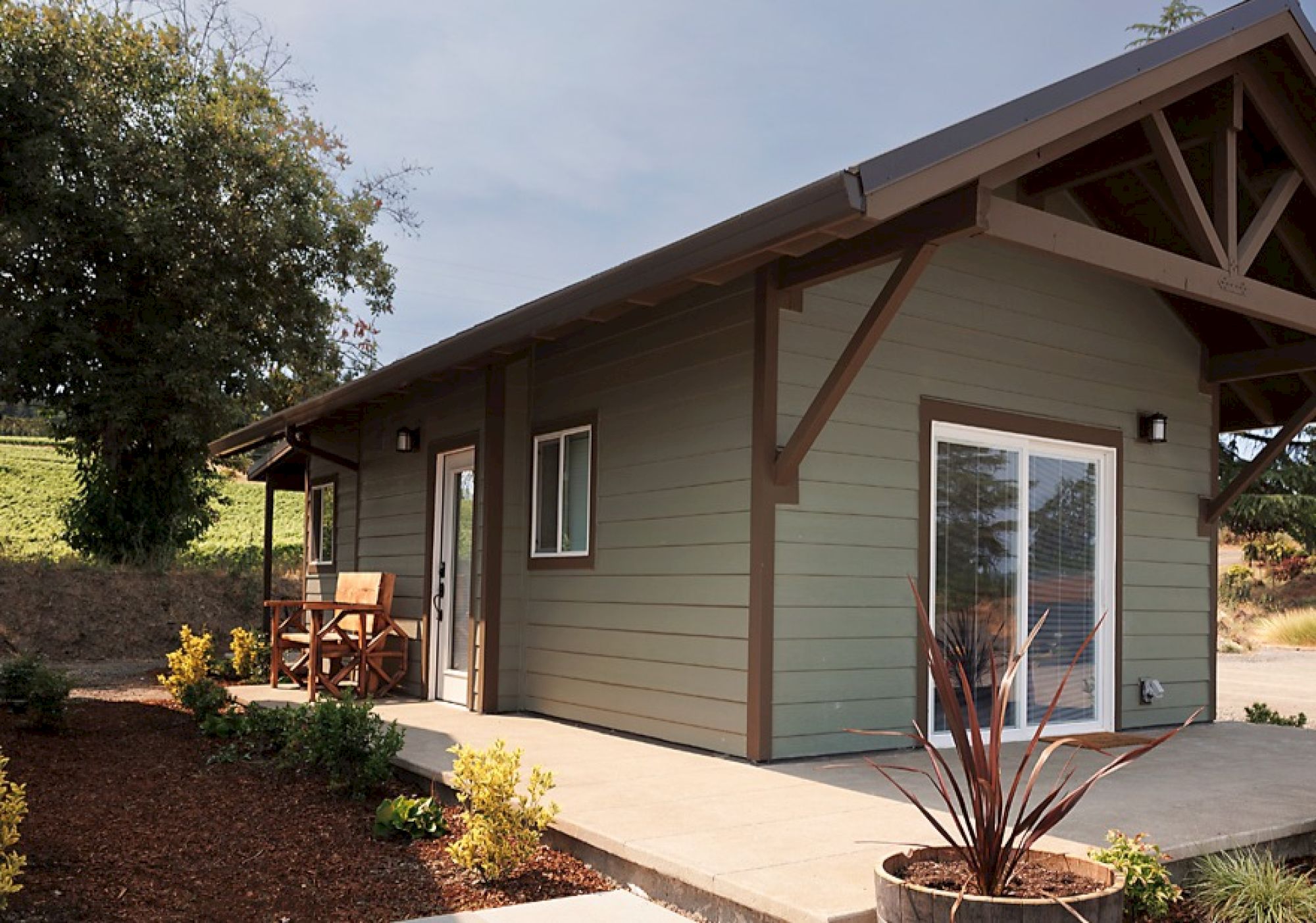 A small, modern house with green siding, a brown roof, patio furniture, and surrounding plants.