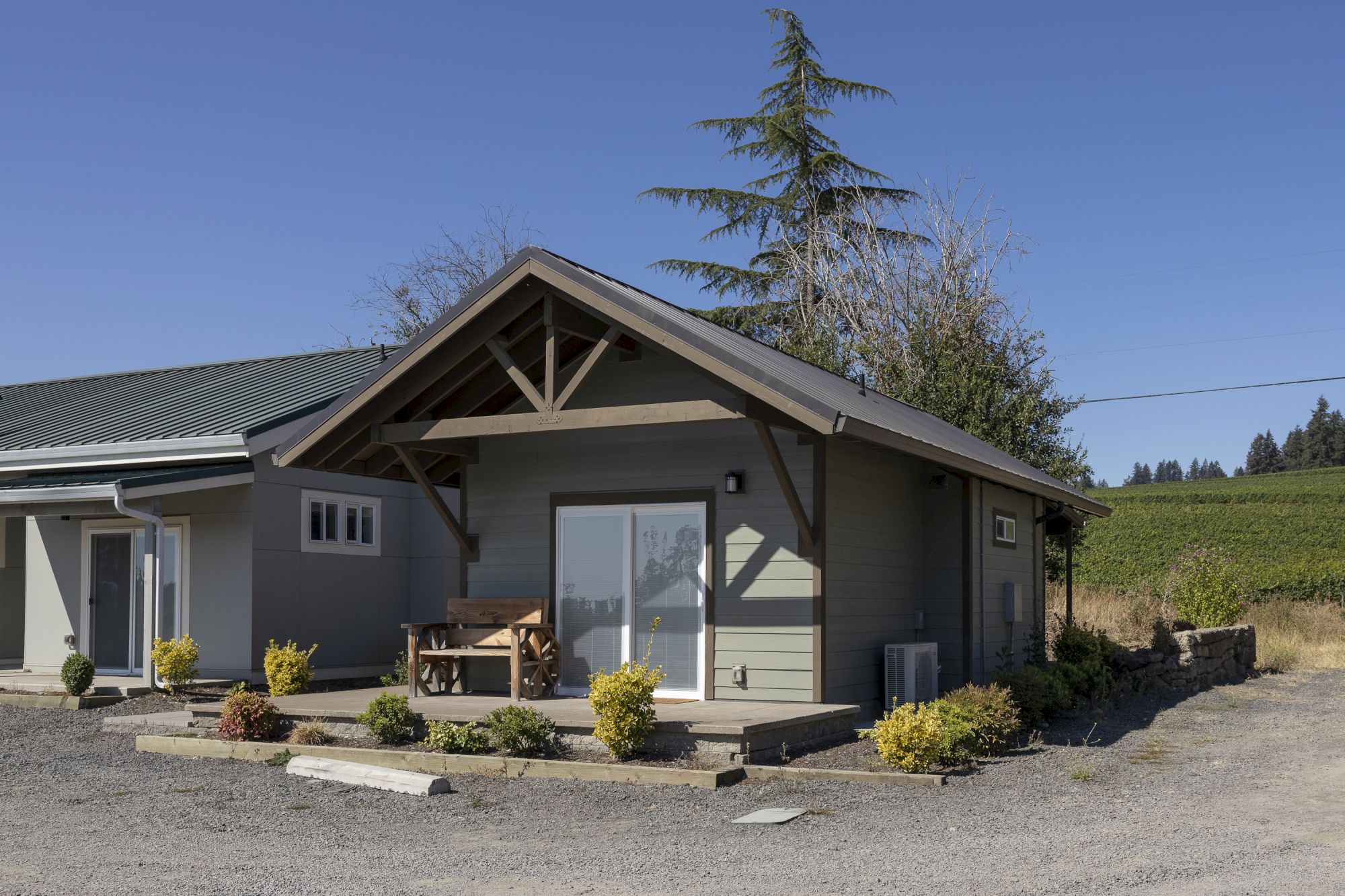 A modern countryside house with a steep gabled roof, large windows, shrubs, and tall trees under a clear blue sky.