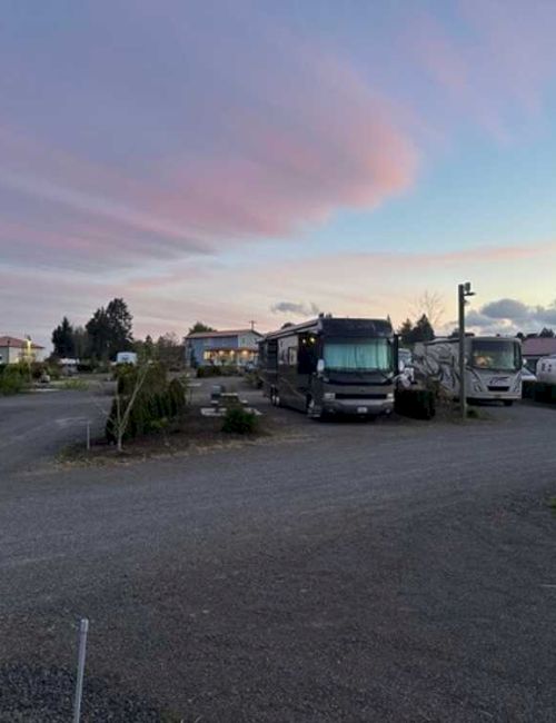 An RV park with motorhomes and trailers, set against a colorful sky at dusk. Trees and picnic tables are visible around the area.