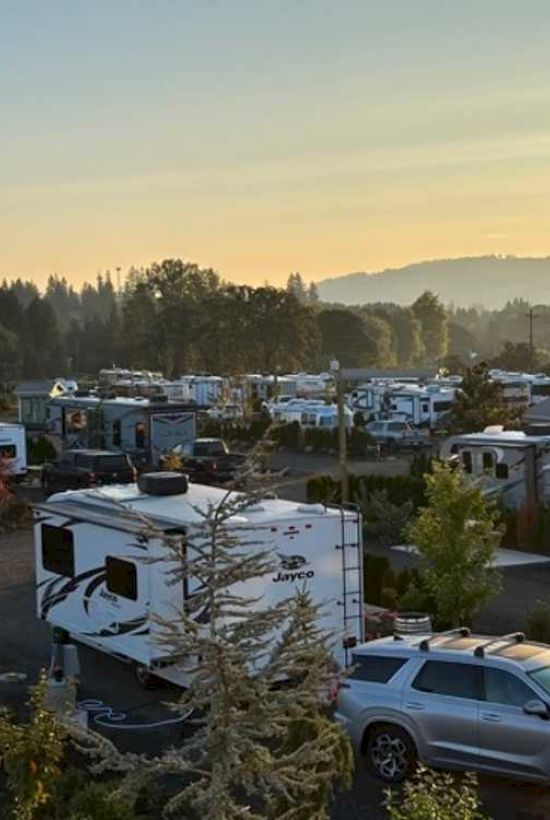 The image shows a campsite with multiple RVs and trailers surrounded by trees, set against a backdrop of hills and a sunrise or sunset.