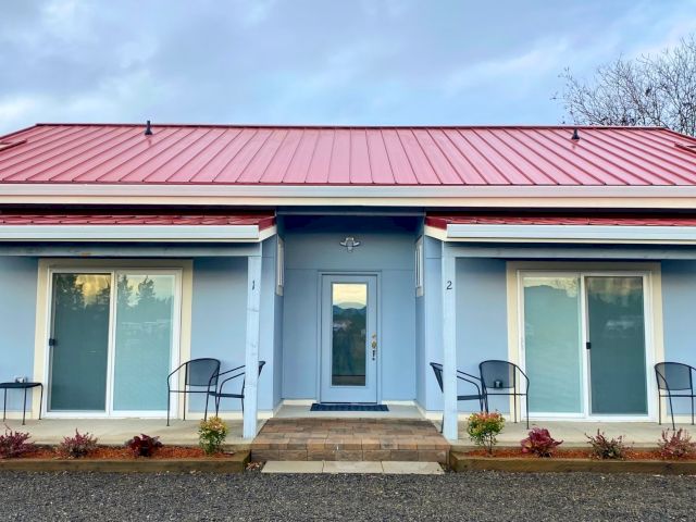 A modern blue duplex with a red roof, sliding glass doors, and porch seating, surrounded by small plants and gravel path in front.