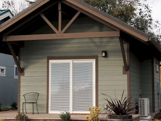 Small house with green siding, sliding glass doors, a chair on the porch, and plants in front.