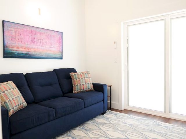 A cozy room with a dark blue sofa, patterned pillows, abstract art on the wall, and large glass doors letting in natural light.