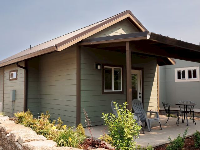 A small house with a covered porch, two chairs, and a table on a patio surrounded by plants and a stone wall.