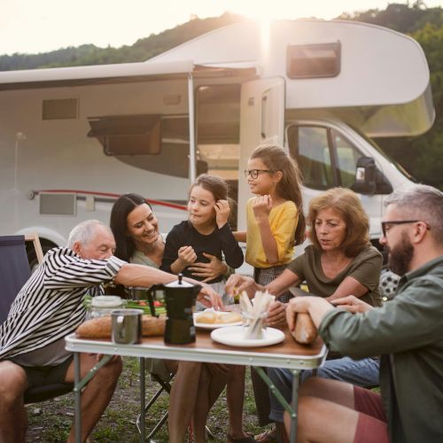 A family enjoys a meal outdoors beside a camper, surrounded by nature. They appear happy and relaxed, sharing food and smiles.