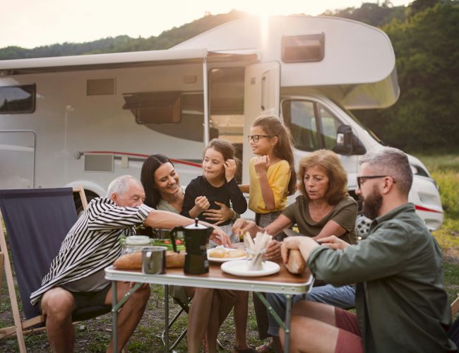 A family enjoys a meal outdoors beside a camper, surrounded by nature. They appear happy and relaxed, sharing food and smiles.