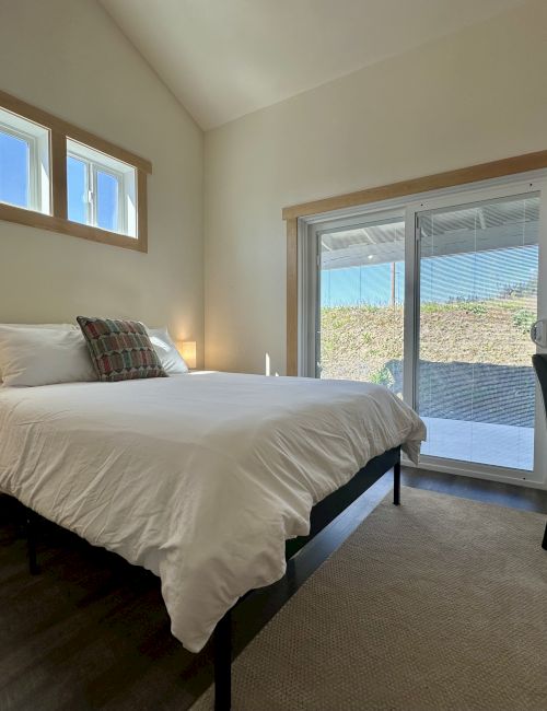 A minimalistic bedroom with a bed, white bedding, two small lamps, chair, and sliding glass doors leading outside.