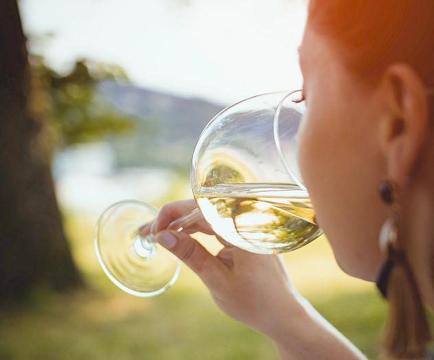 A person is outdoors, drinking from a glass of white wine, with green grass and a tree in the background.