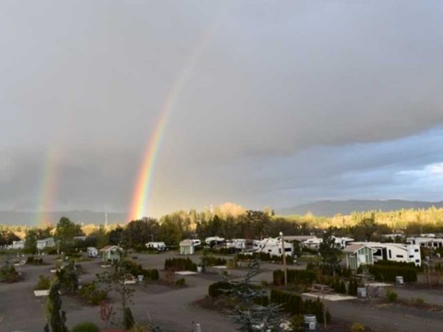 The image shows a landscape with a double rainbow over a campground with several RVs, surrounded by trees and mountains in the background.