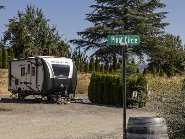A camper trailer is parked near a green sign that says &ldquo;Pinot Circle,&rdquo; with trees and a paved road around it.