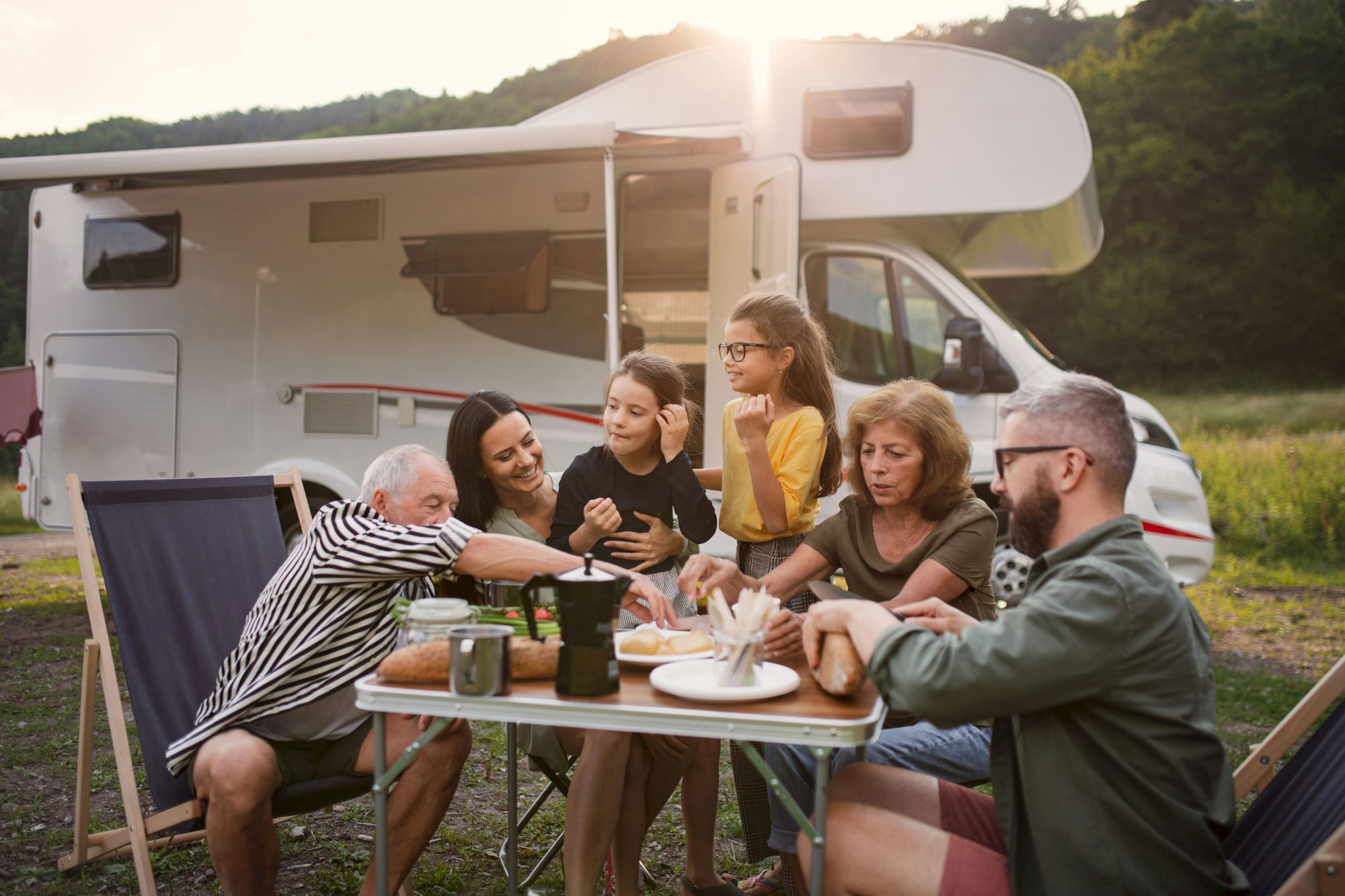 A group enjoys a meal outdoors by a camper van, surrounded by trees and hills, with the sun setting in the background.