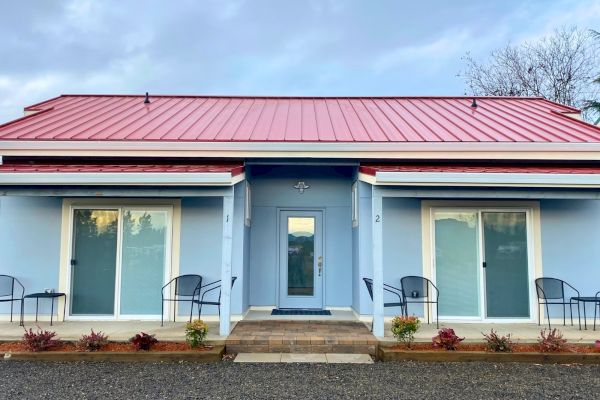 A blue building with a red roof features two sliding glass doors and small seating areas, surrounded by plants at the front.