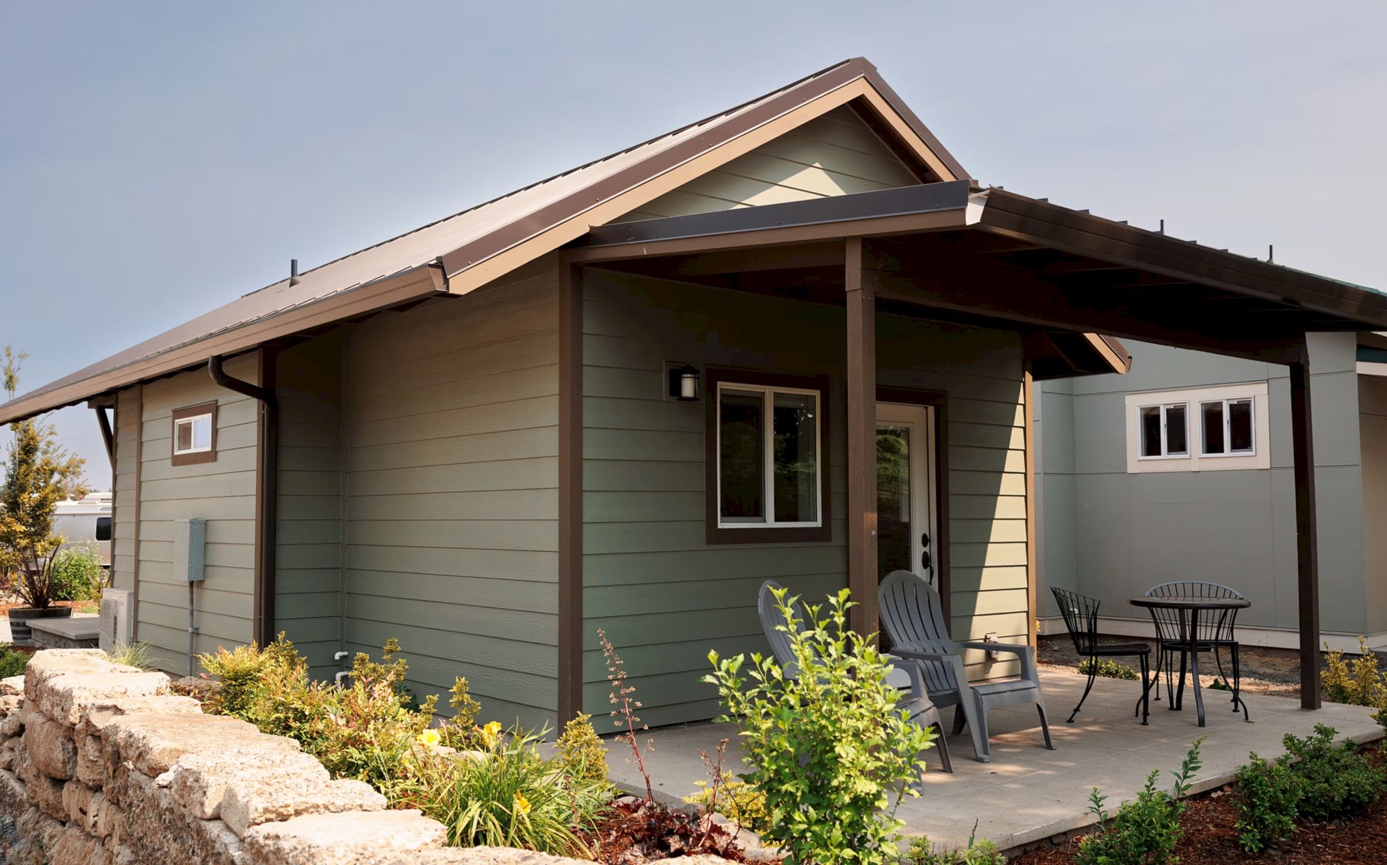 A small house with a covered porch, chairs, and a table, surrounded by plants under a clear sky.