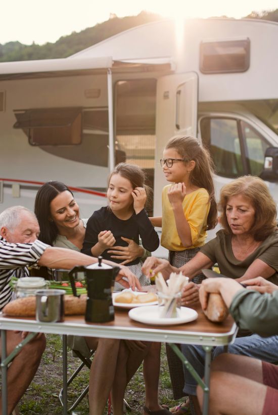 A family enjoys a meal outdoors near a campervan, surrounded by nature and sunshine, sharing food and laughter at a table.