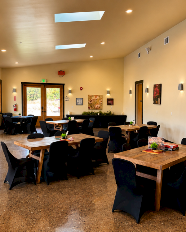 A modern community hall with wooden tables and black chairs, wall art, large doors, and warm lighting fills the room.