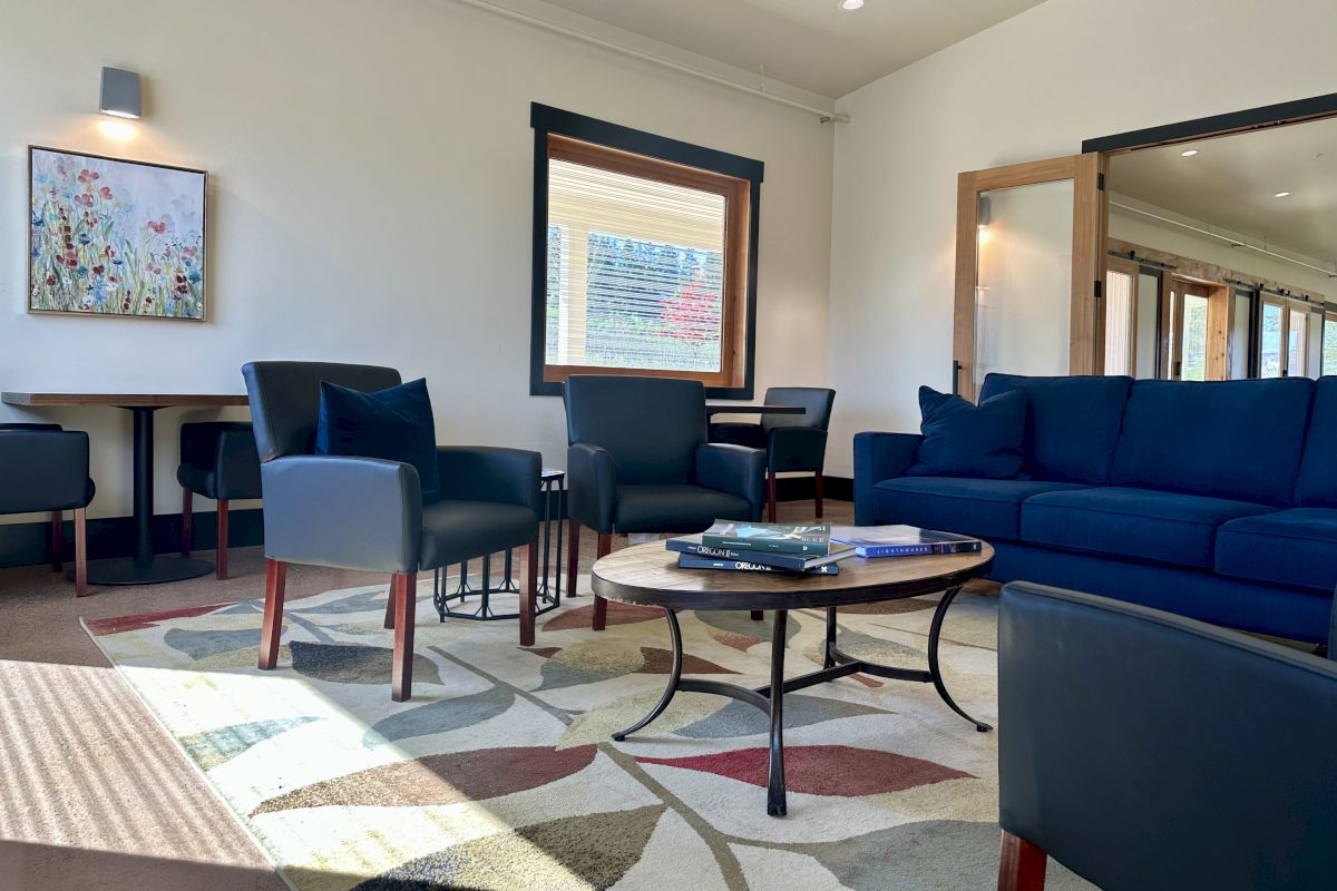 A bright living room with blue sofas and chairs, a round coffee table with books, and large windows letting in daylight.