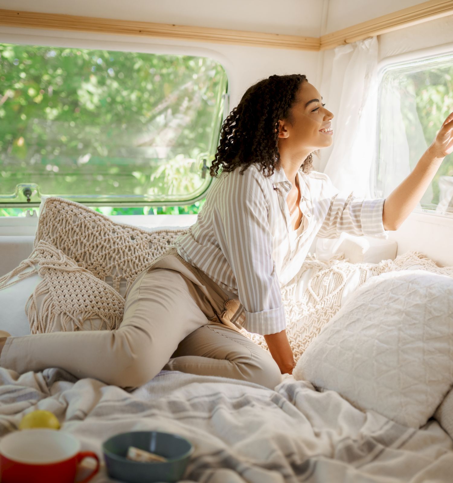 A person is relaxing on a bed inside a cozy space, looking out the window. There are pillows and a cup nearby.