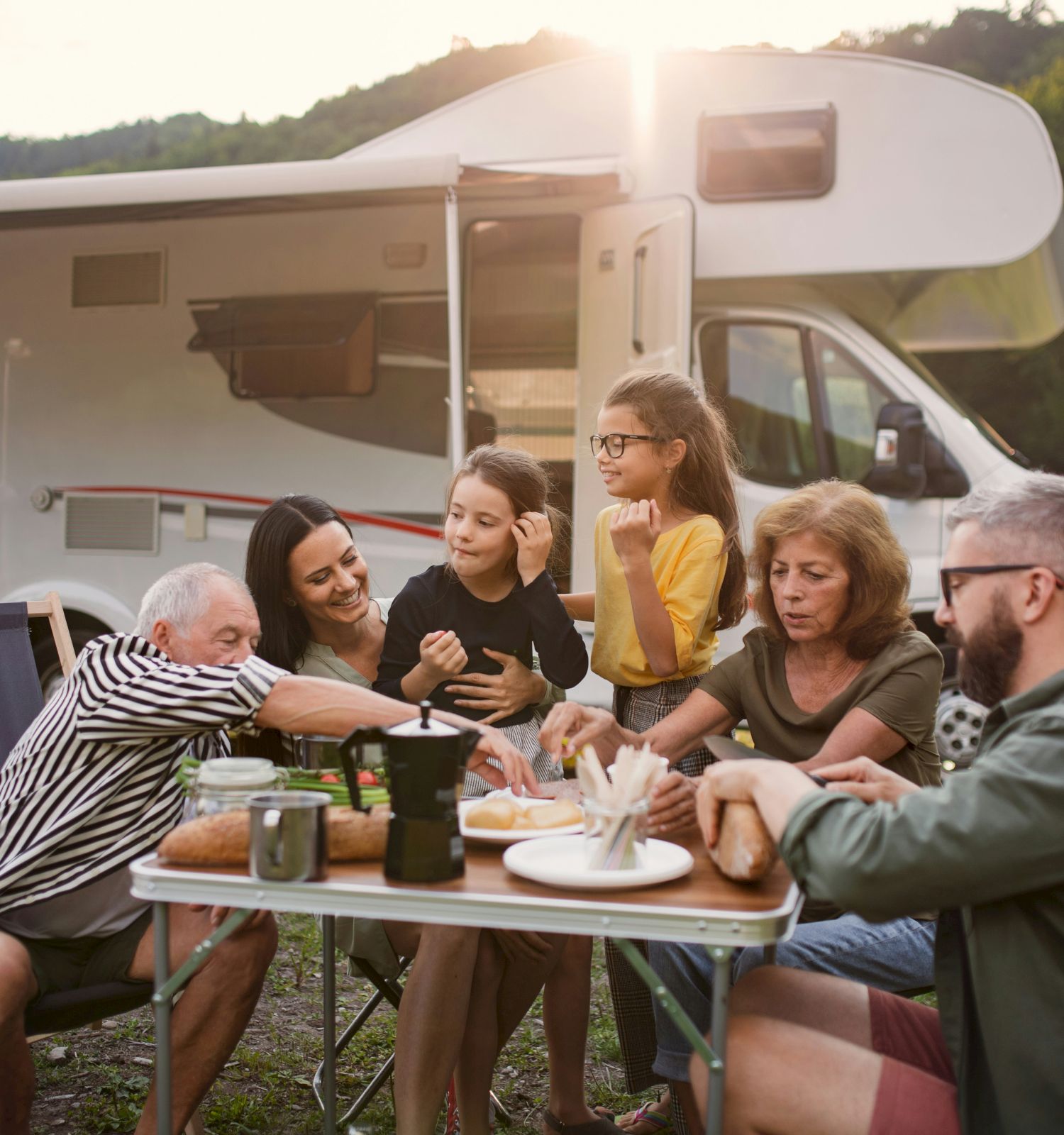 A group of people are enjoying a meal together outdoors next to a camper, surrounded by nature, with the sun setting behind them.