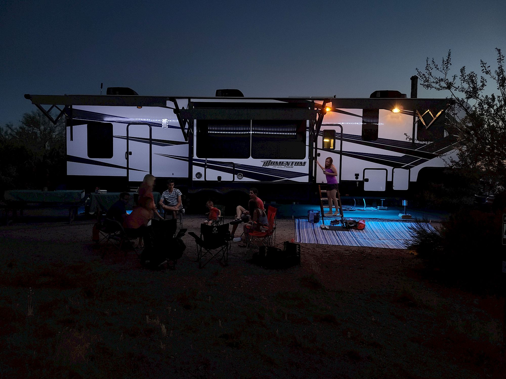A group of people is gathered outside a large RV at dusk, with one person on a ladder, and soft lights illuminate the scene.