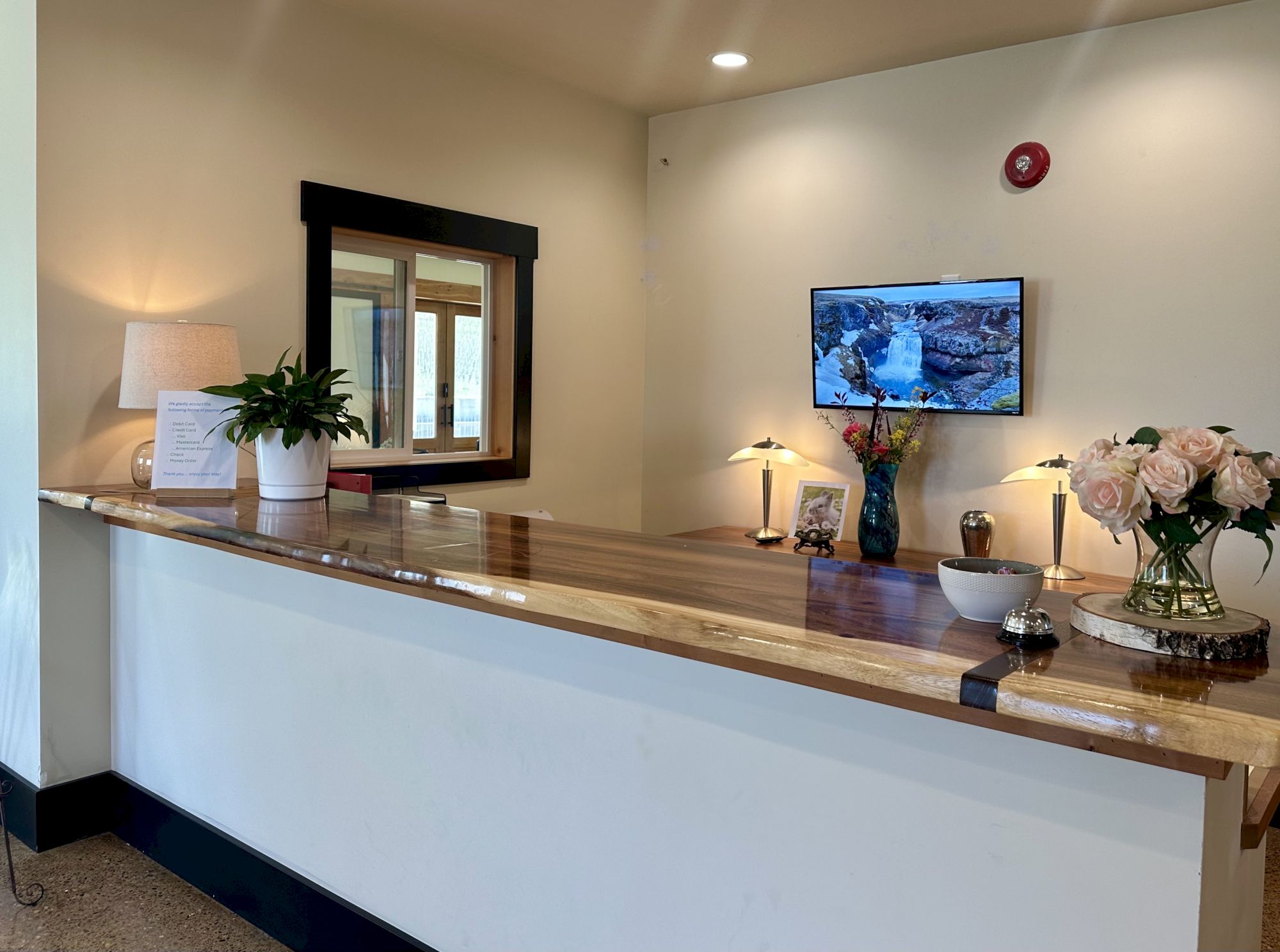 A reception desk with a lamp, flowers, framed pictures, and a TV on the wall displaying a waterfall scene.