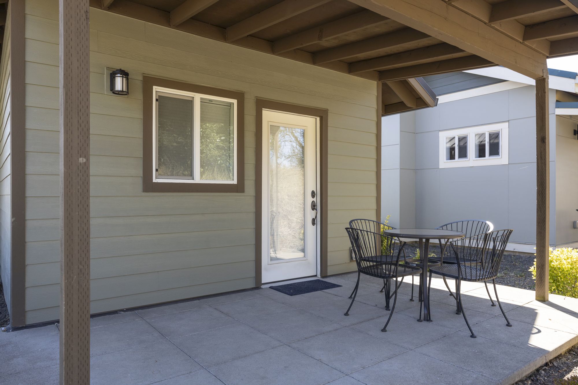 Covered patio with beige siding, a front door and windows, and a black metal table with chairs for outdoor dining.