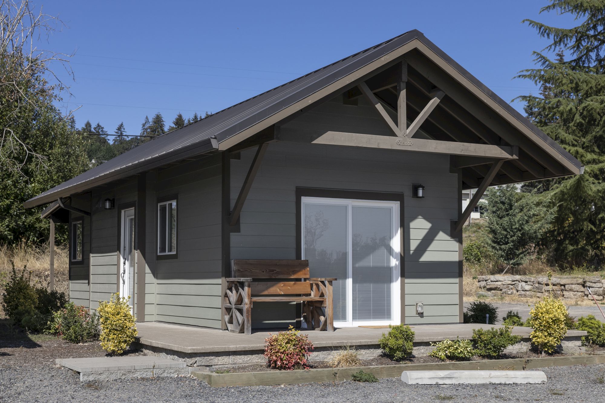 A modern gray house exterior with a steep metal roof, front porch overhang, windows, and a wooden bench on a sunny day.